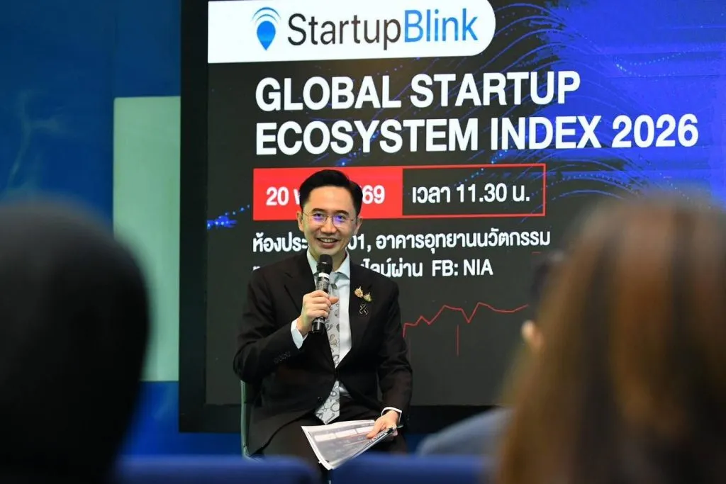 The same man sits on stage, smiling warmly while holding a microphone and papers. The screen behind displays startup index branding and Thai text.