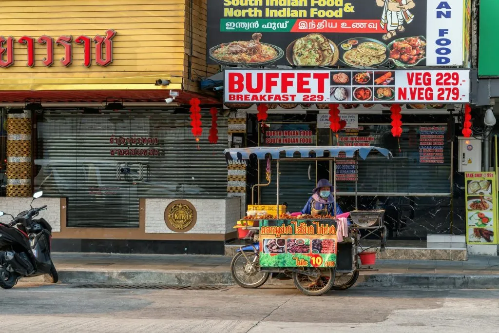 A masked street vendor sits at a motorized food cart displaying meat skewers, parked beneath brightly colored restaurant signs advertising Indian buffets.