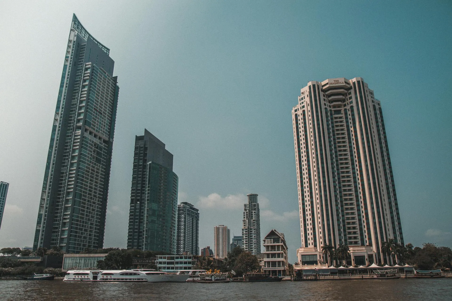 Tall, modern skyscrapers loom over a wide river under a clear sky. Several passenger boats are moored along the water's edge in the foreground.