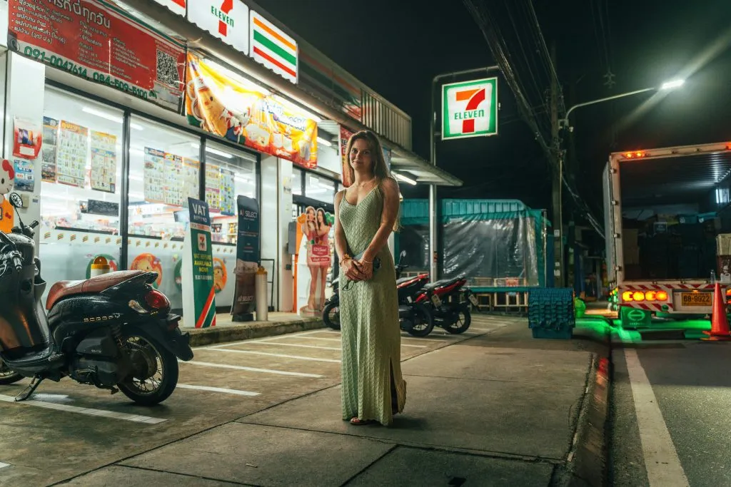 A woman in a sage dress stands before a glowing 7-Eleven at night, surrounded by parked scooters and a delivery truck.