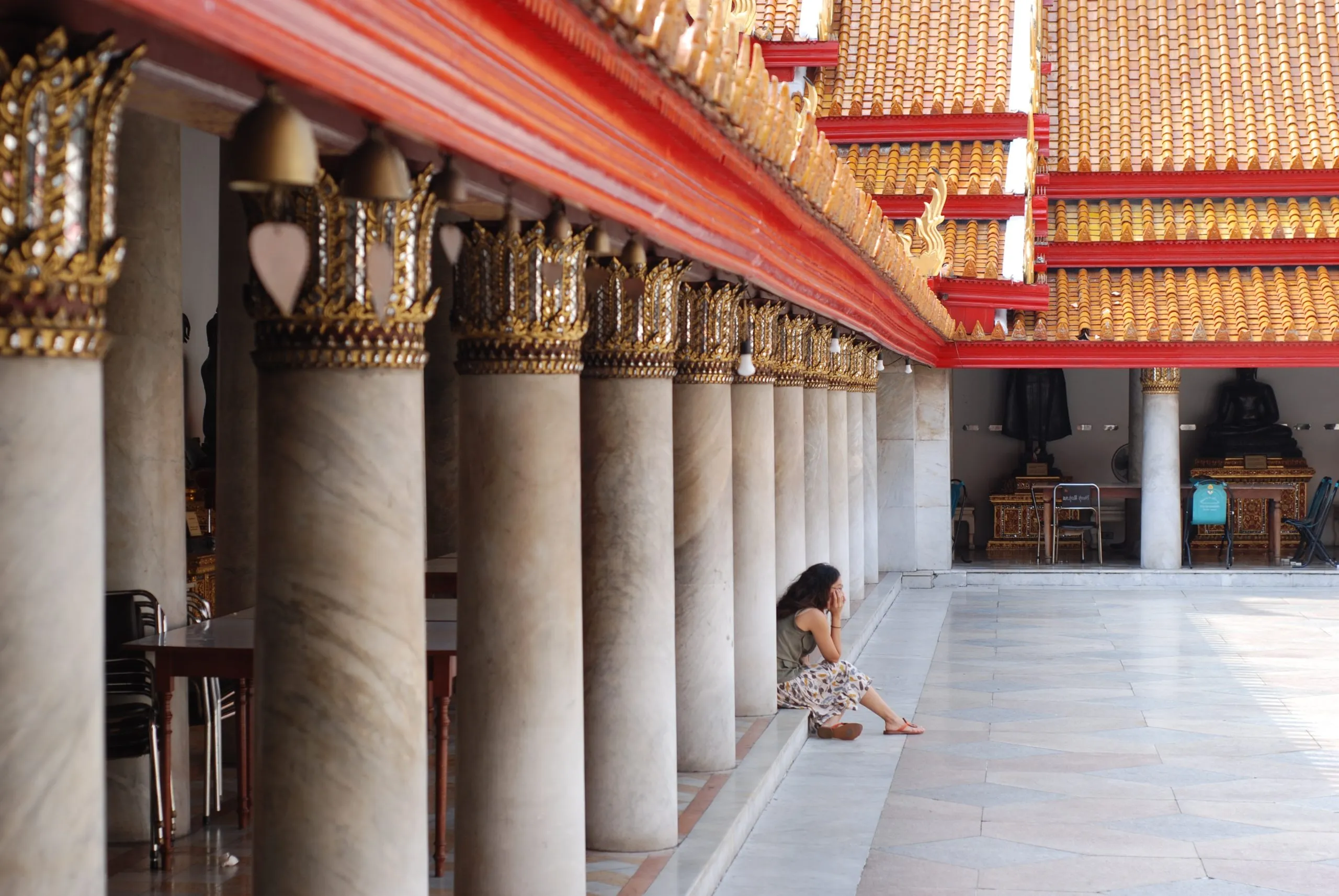 A woman sits, resting against a row of thick marble pillars under the ornate red and gold eaves of a tranquil Thai temple courtyard.