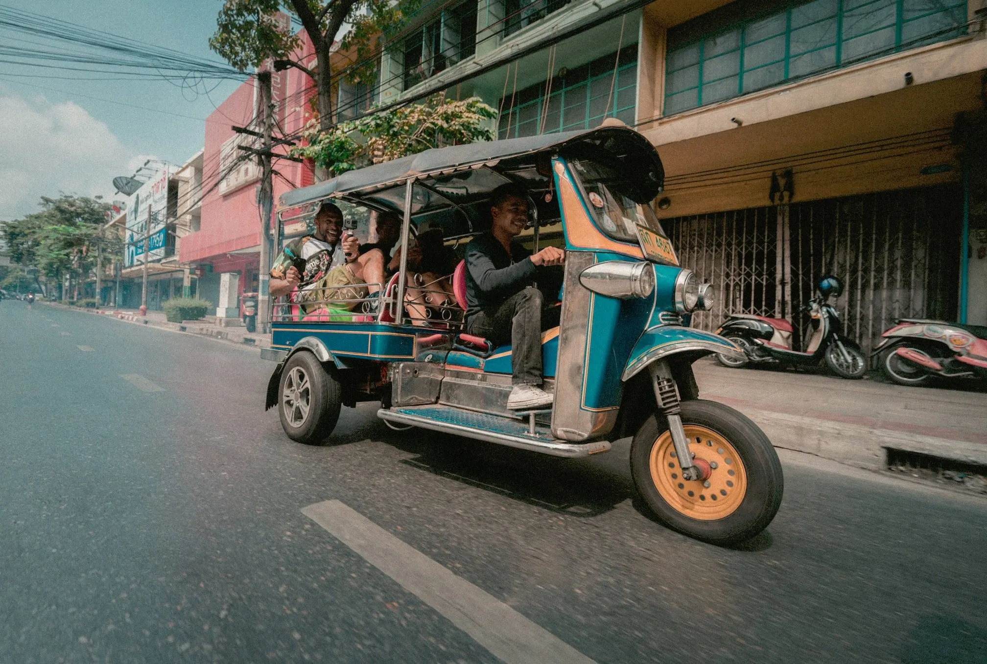 A blue and yellow tuk-tuk carries smiling passengers past city buildings.