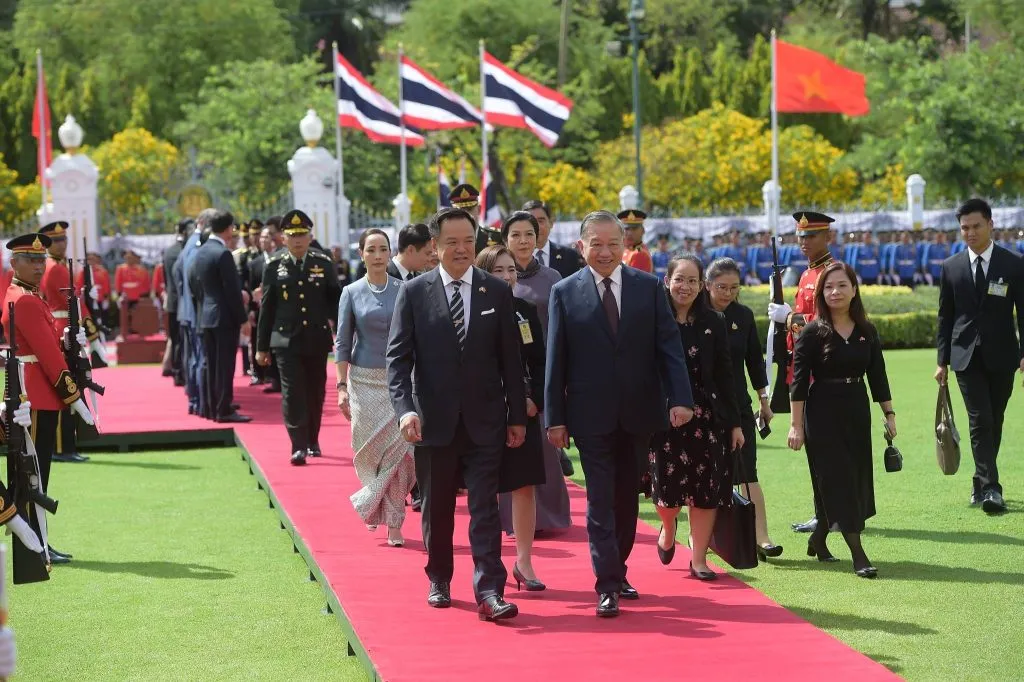 Two men in suits walk along an outdoor red carpet, followed by a large group.