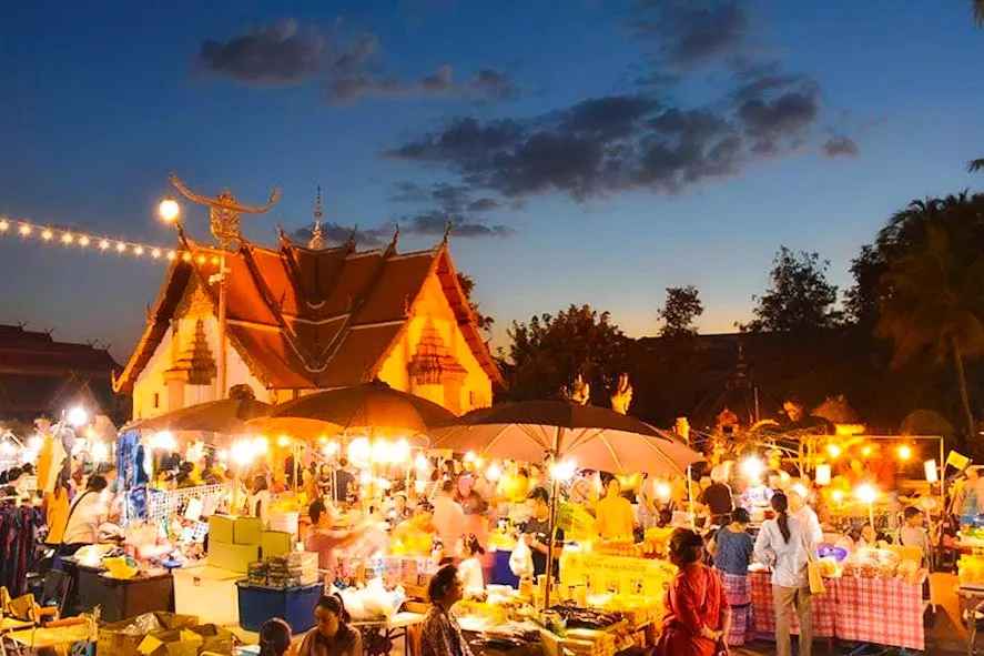 A bustling night market glowing with warm lights under a twilight sky, set against the illuminated backdrop of Wat Phumin's distinctive multi-tiered roofs.
