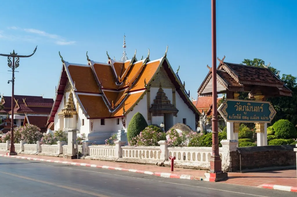 A sunny street-side view of Wat Phumin in Nan, Thailand, featuring an ornate white temple with multi-tiered orange roofs behind a low white fence.