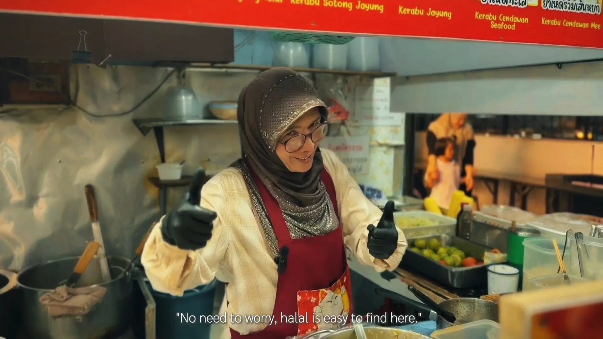 A woman wearing a brown hijab and red apron stands in a kitchen, smiling and giving two thumbs up while wearing black protective gloves.