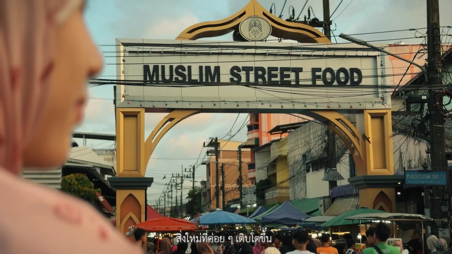 A large golden archway spans a busy street market, displaying bold black letters reading MUSLIM STREET FOOD against a background of urban utility lines.