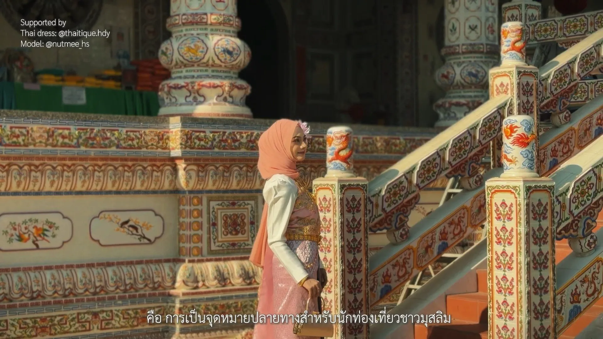 A woman in a pink hijab and traditional Thai dress stands before an ornately decorated temple structure featuring colorful mosaic pillars and a dragon motif.