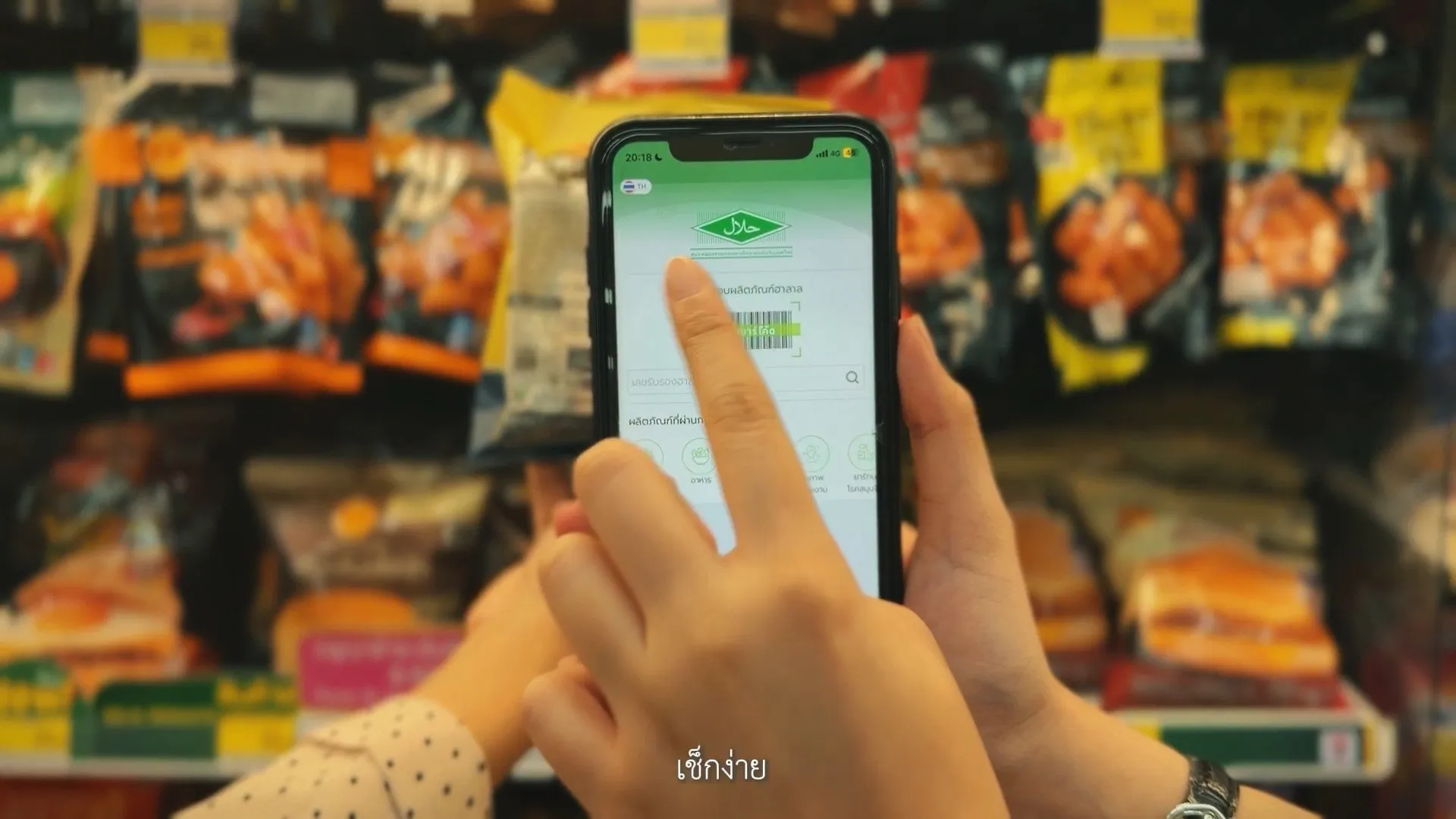 Hands hold a smartphone displaying a green halal certification app with a barcode, positioned in front of blurred grocery store shelves filled with various snacks.