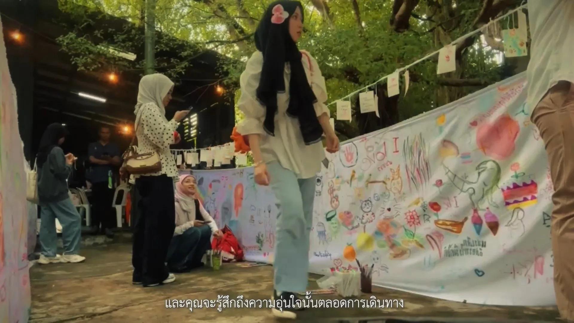 Young women in hijabs walk past a large outdoor white banner covered in colorful, hand-drawn childlike illustrations and paintings under a canopy of trees.