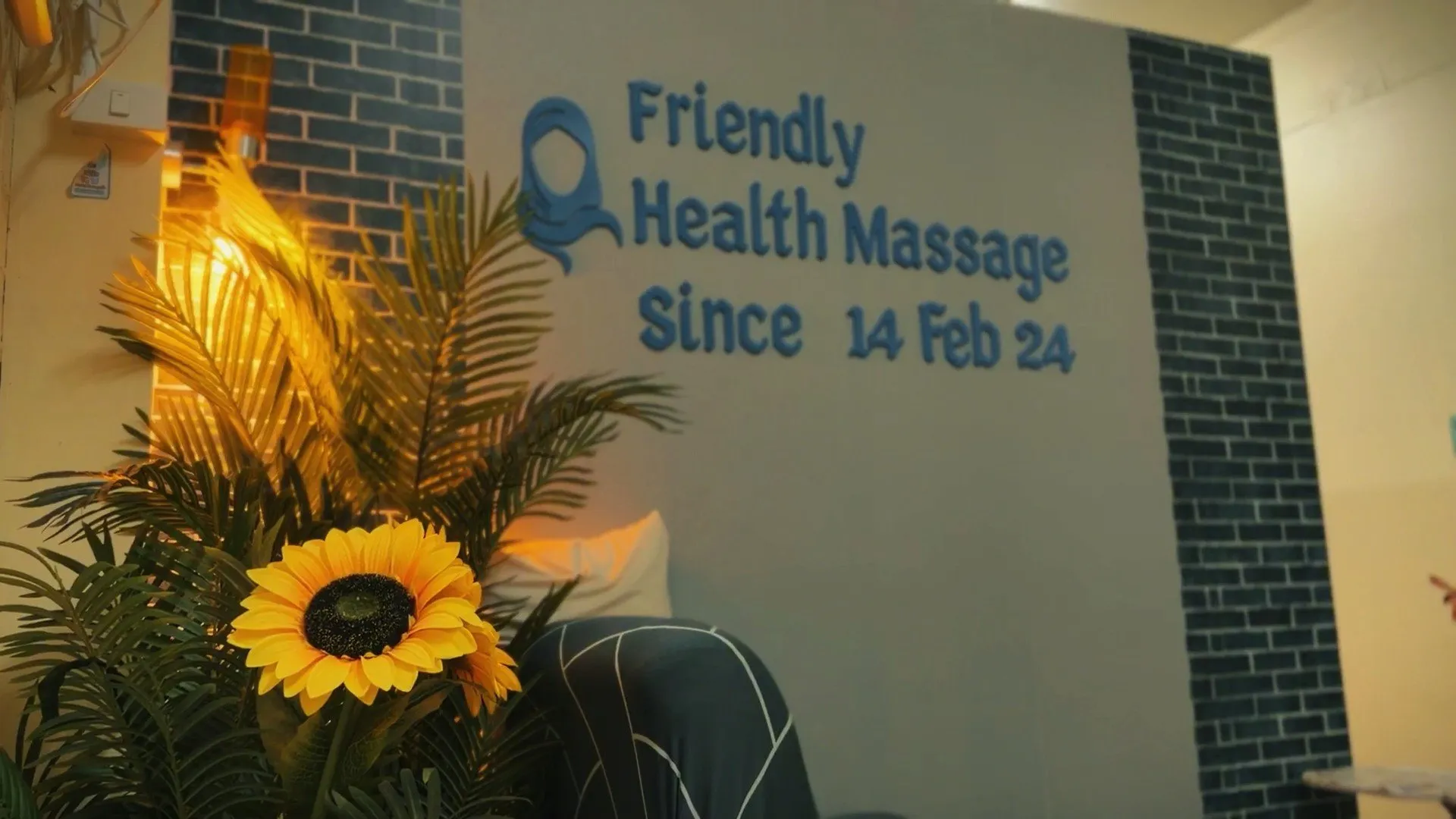 A gray wall features blue lettering reading Friendly Health Massage Since 14 Feb 24, accompanied by a decorative sunflower and a blue hijab icon.