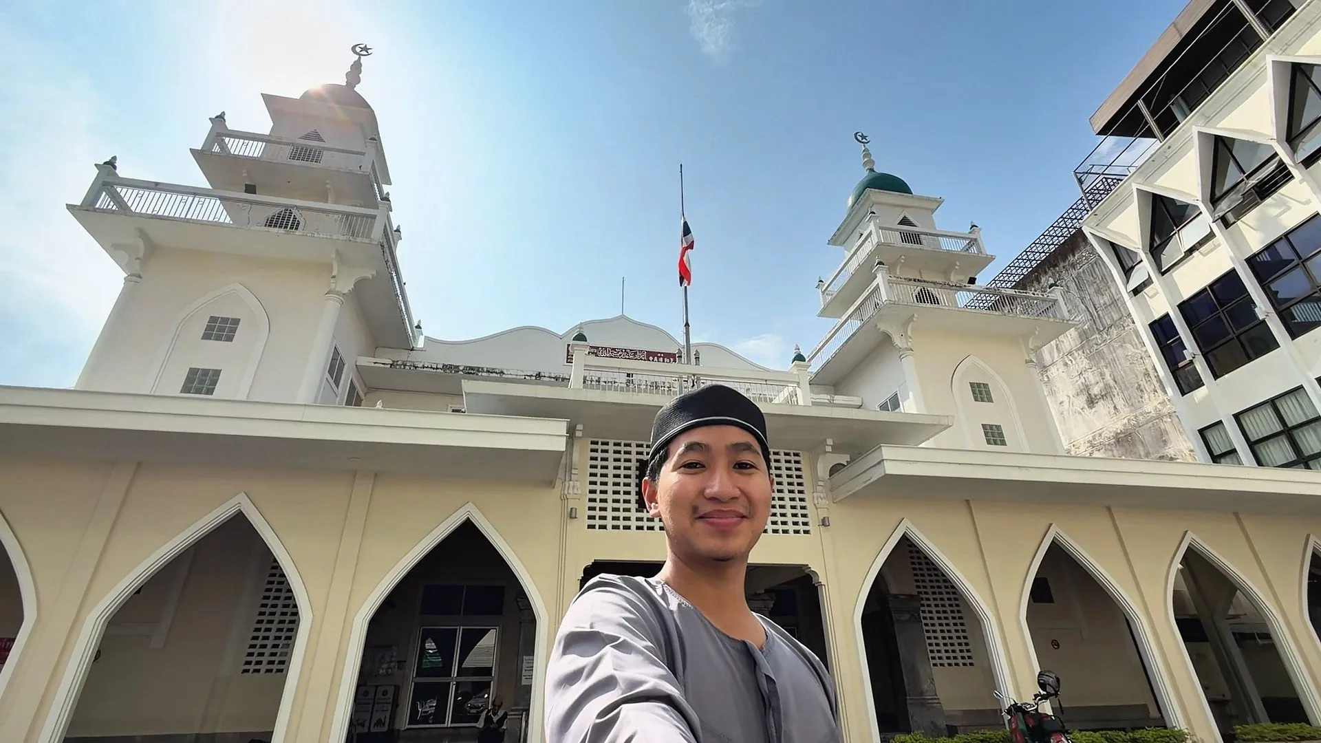 A man in a grey tunic and black prayer cap smiles for a selfie in front of a white mosque with pointed arches and minarets.