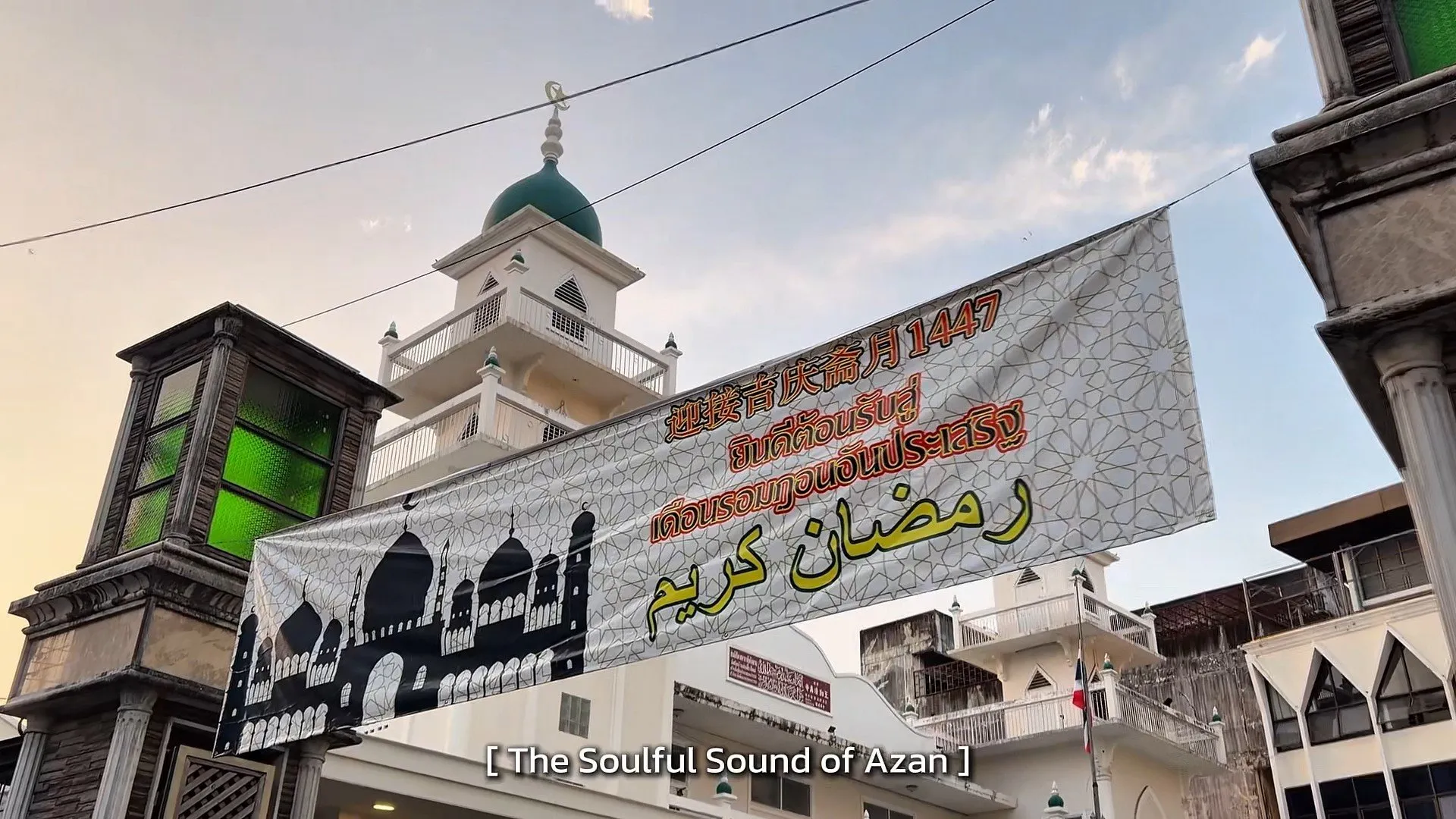 A white banner with colorful Thai and Arabic script welcoming the holy month of Ramadan hangs between buildings, with a mosque minaret in the background.