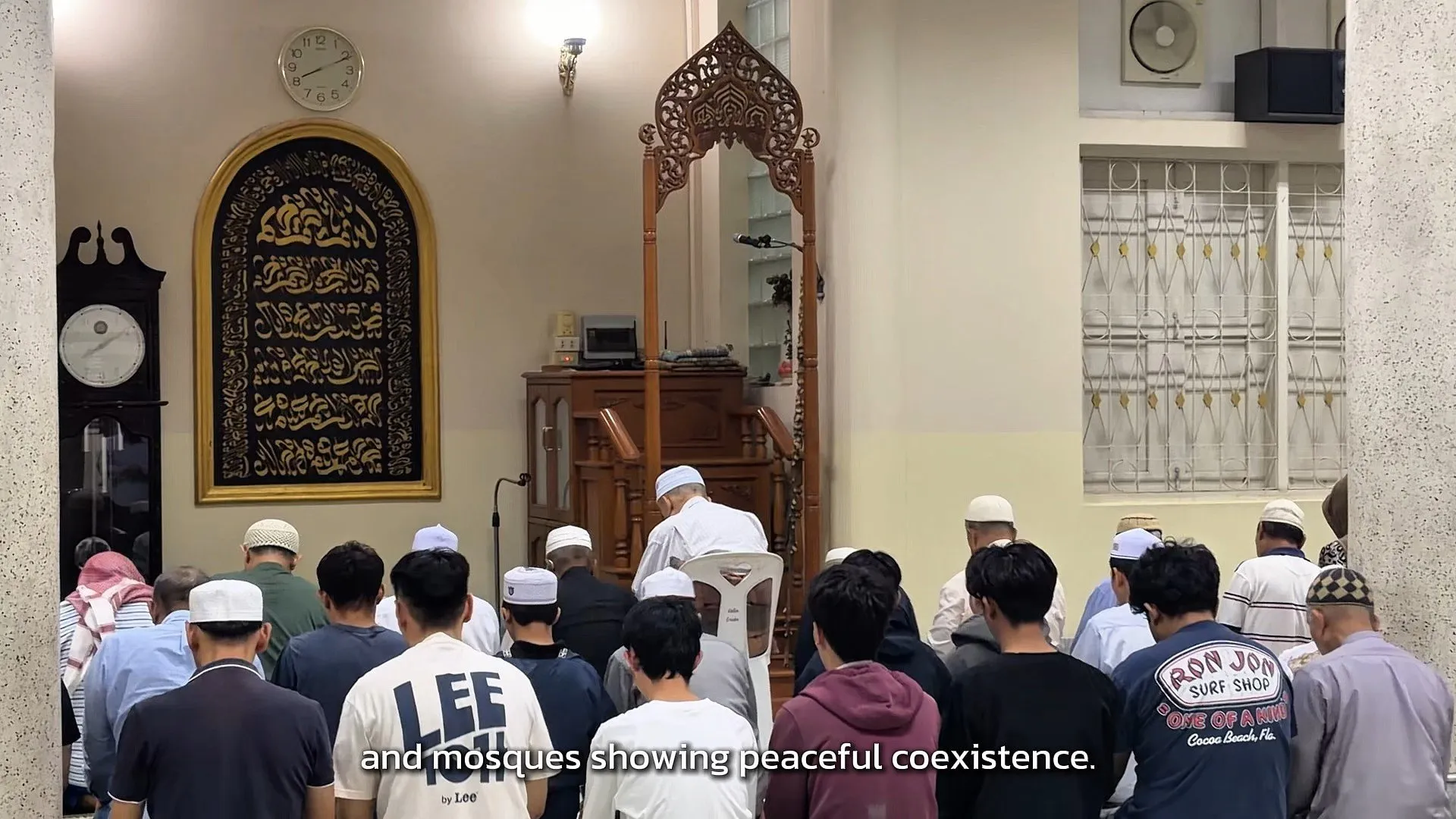 Inside a prayer hall, a group of men wearing caps sit on the floor facing a decorative wooden minbar and gold-framed calligraphy on the wall.