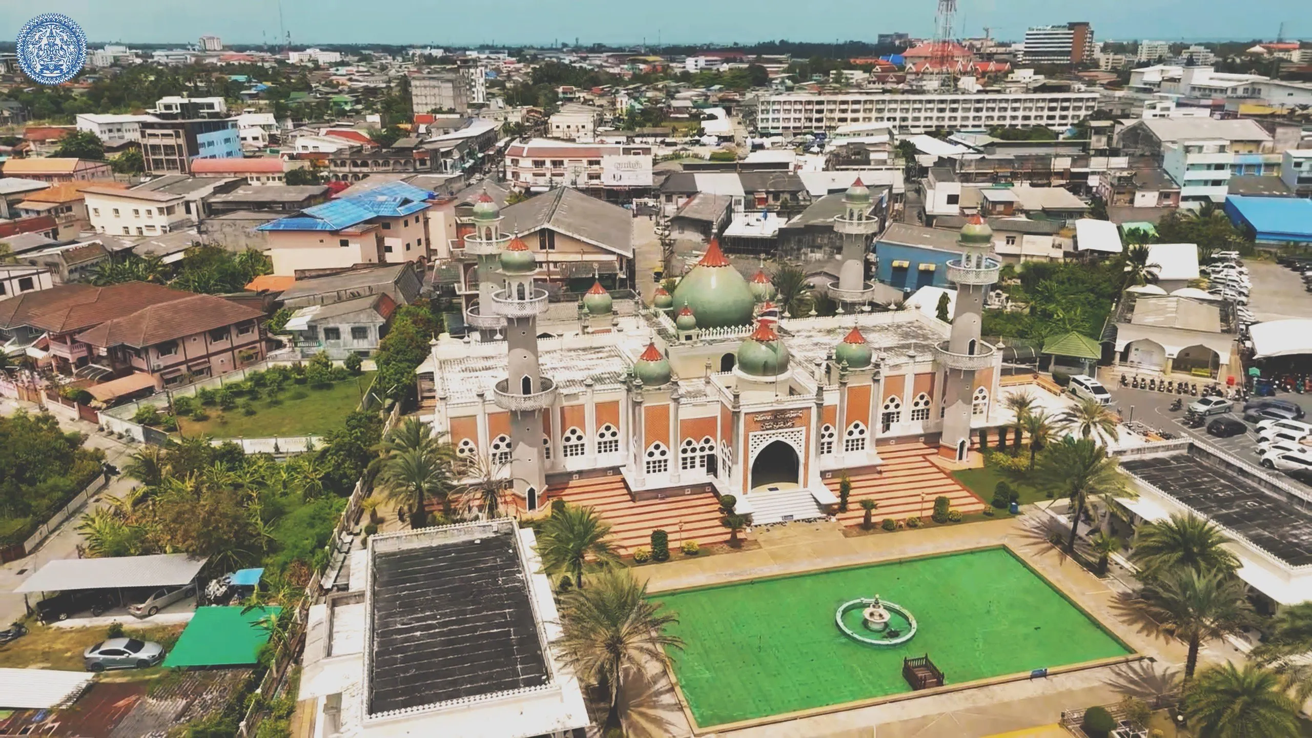 High-angle view showing a majestic mosque with green domes and minarets, surrounded by a pond and an urban landscape under a bright, clear sky.