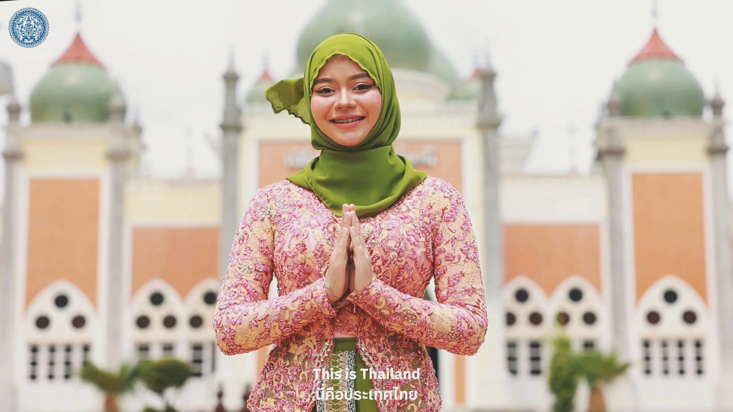 A smiling woman wearing a vibrant green hijab and traditional pink lace top poses with palms pressed together before a grand, mosque-like background.
