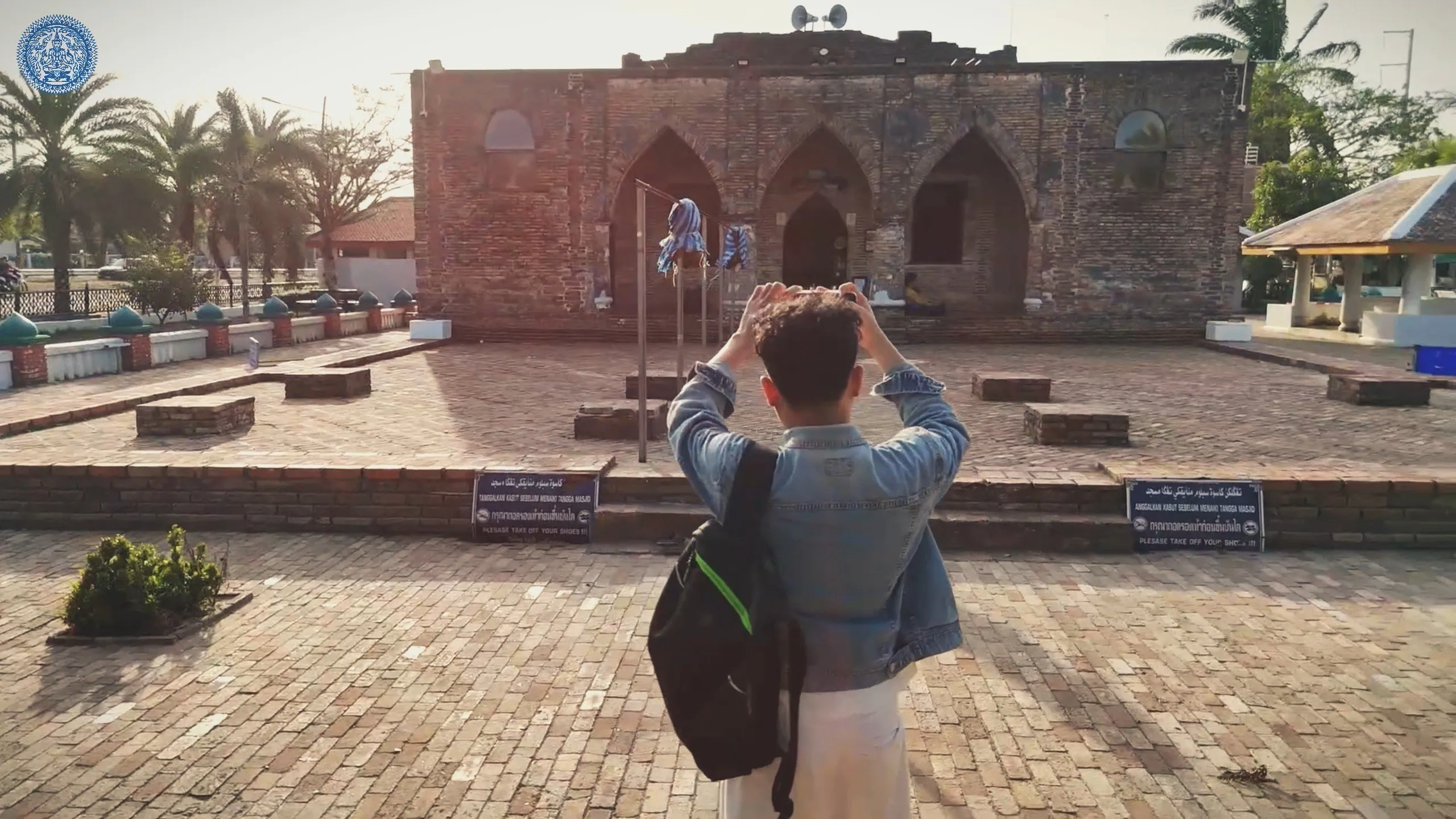 A man in a denim jacket stands with his back to us, photographing an ancient, weathered brick mosque featuring pointed arches and historic character.