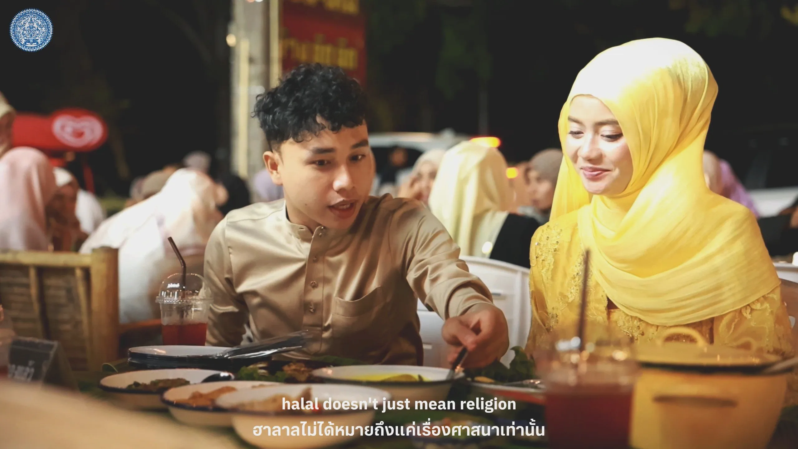 A man and a woman in a yellow hijab sit at a crowded outdoor table, sharing a flavorful meal in a warm nighttime atmosphere.