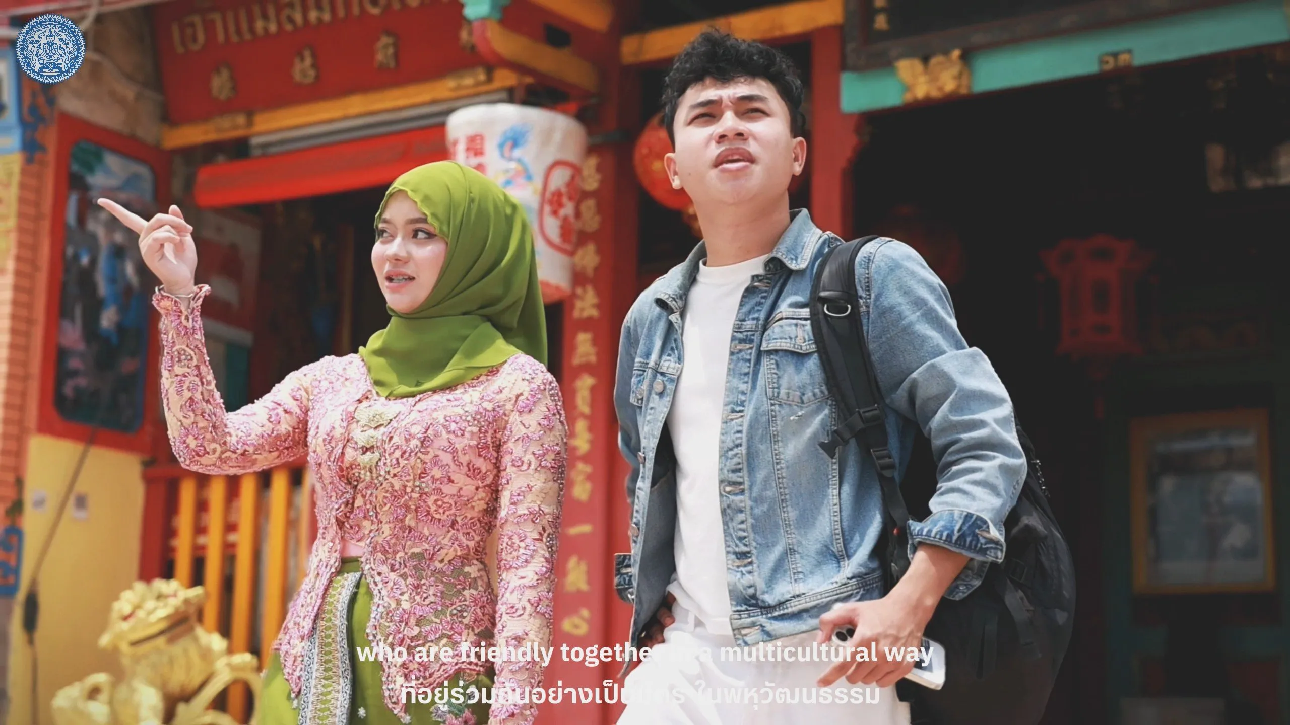 A woman in a green hijab points toward a red Chinese shrine while a man with a backpack observes, showcasing a harmonious multicultural environment.