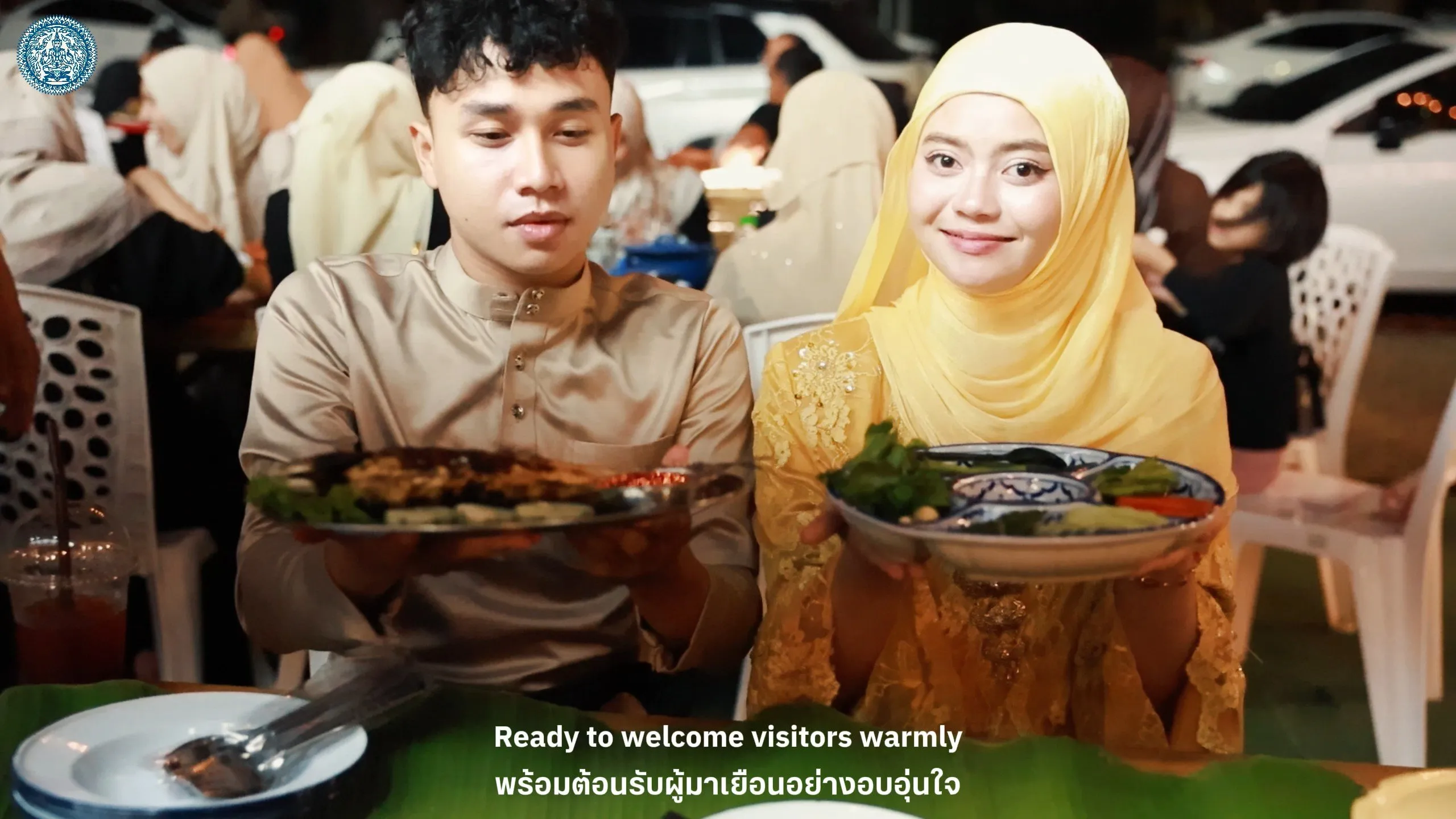 A couple in traditional attire smiles while presenting large plates of food to the camera, inviting visitors to experience local hospitality at night.