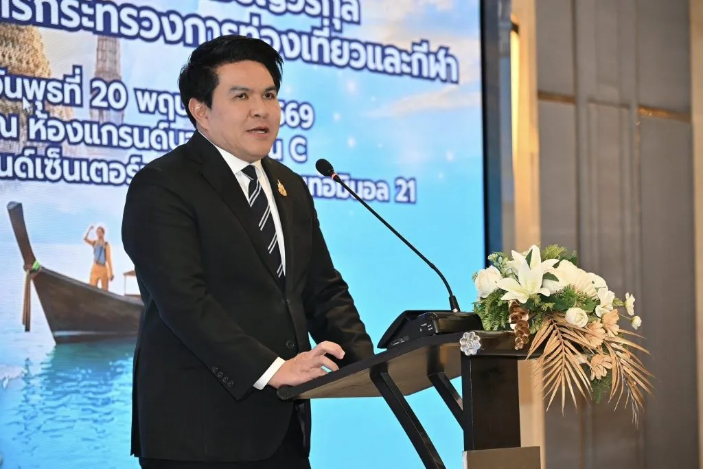 A closer view of the suited man speaking at the floral podium. The brightly lit screen behind him displays a faint image of a boat.