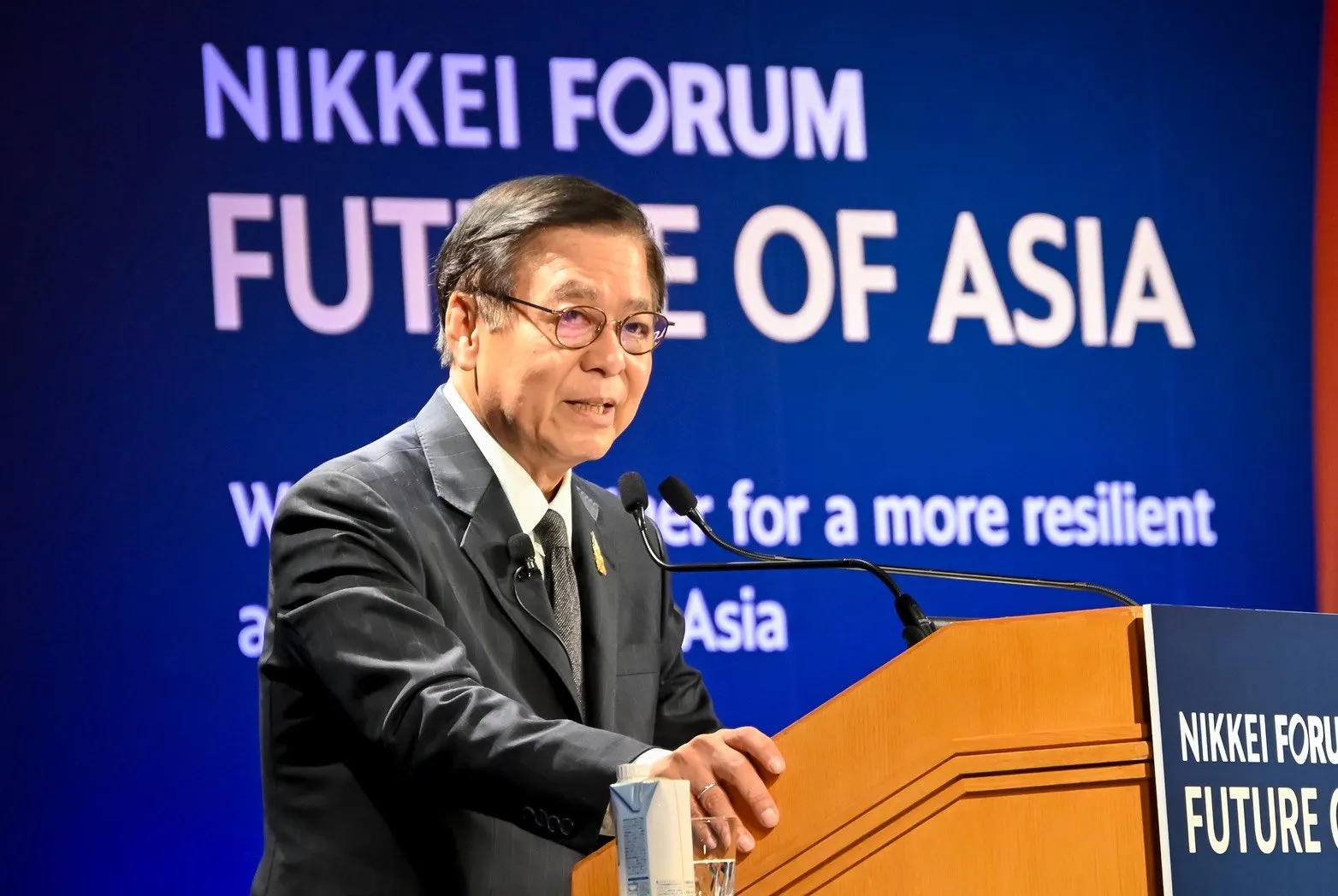 A man in a suit speaks at a podium in front of a blue forum backdrop.