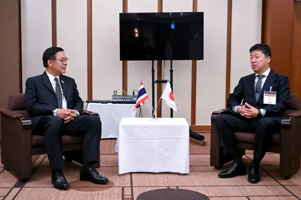 Two men in suits sit facing each other with miniature Thai and Japanese flags between them.