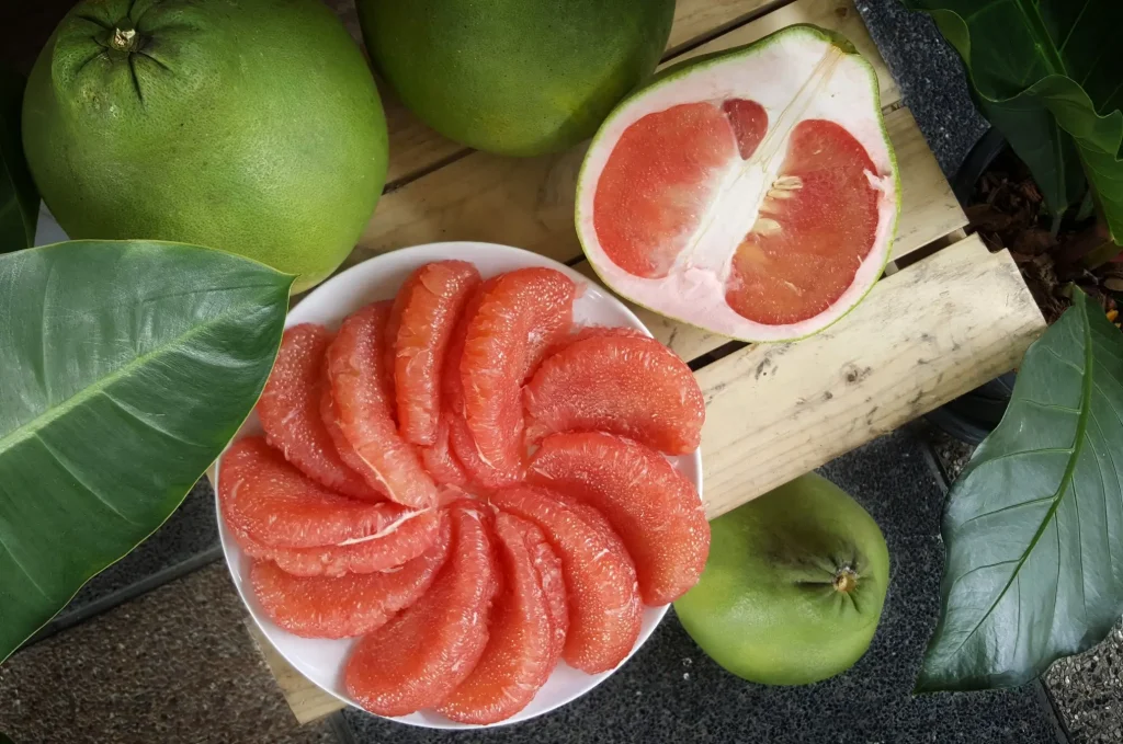 A round plate filled with red, peeled citrus wedges, surrounded by whole green fruits.