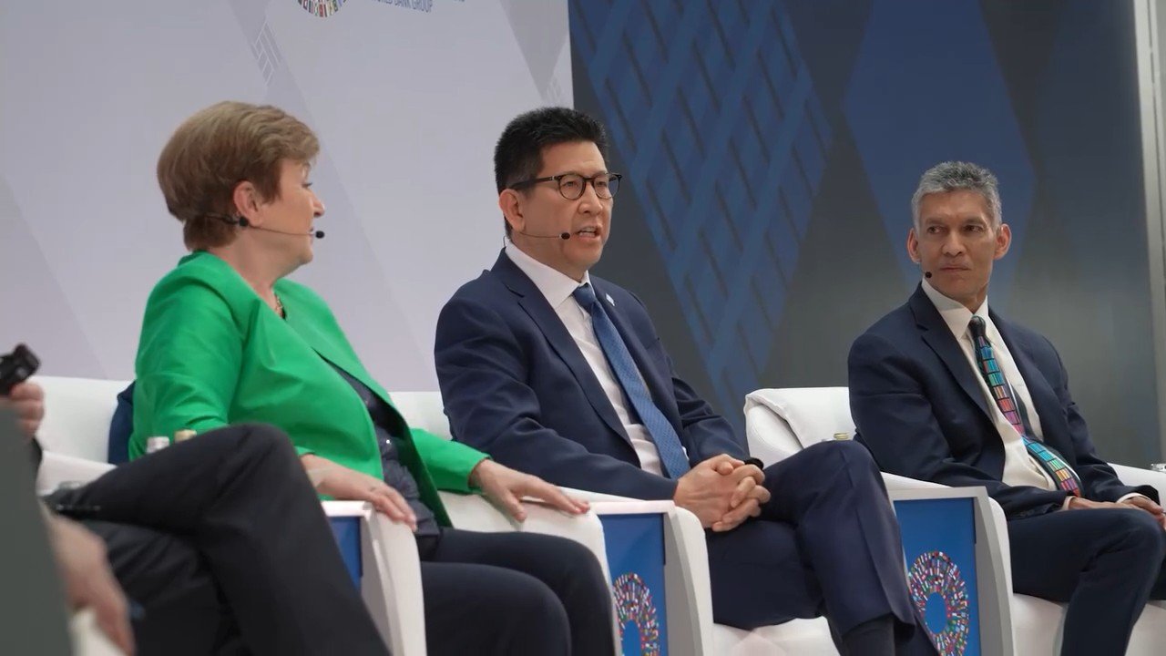 Three panelists in business suits sit on stage during a formal discussion event.