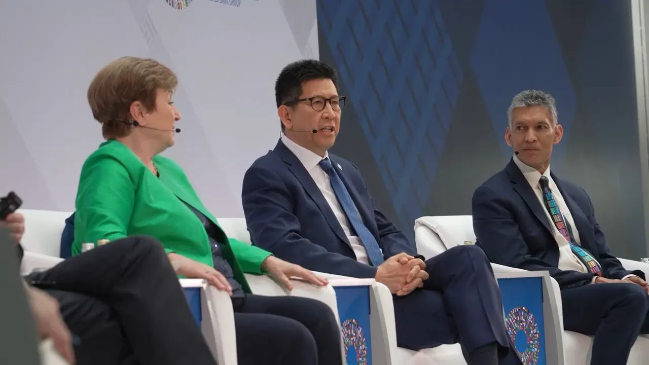 Three panelists in business suits sit on stage during a formal discussion event.