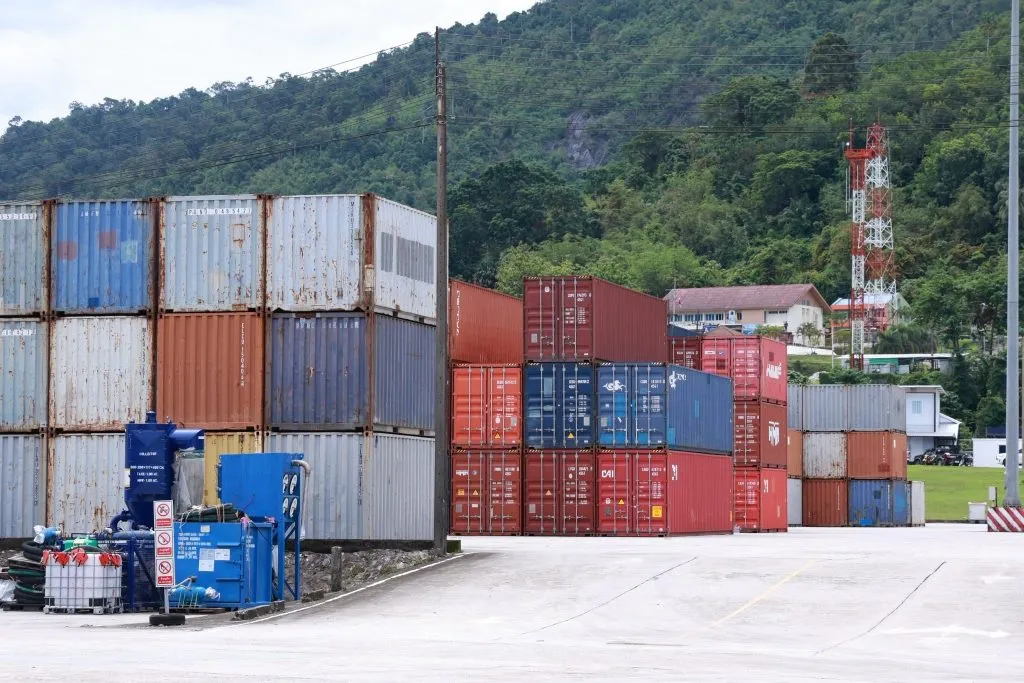 Stacks of colorful, weathered shipping containers sit on a paved lot against a lush, forested hill.