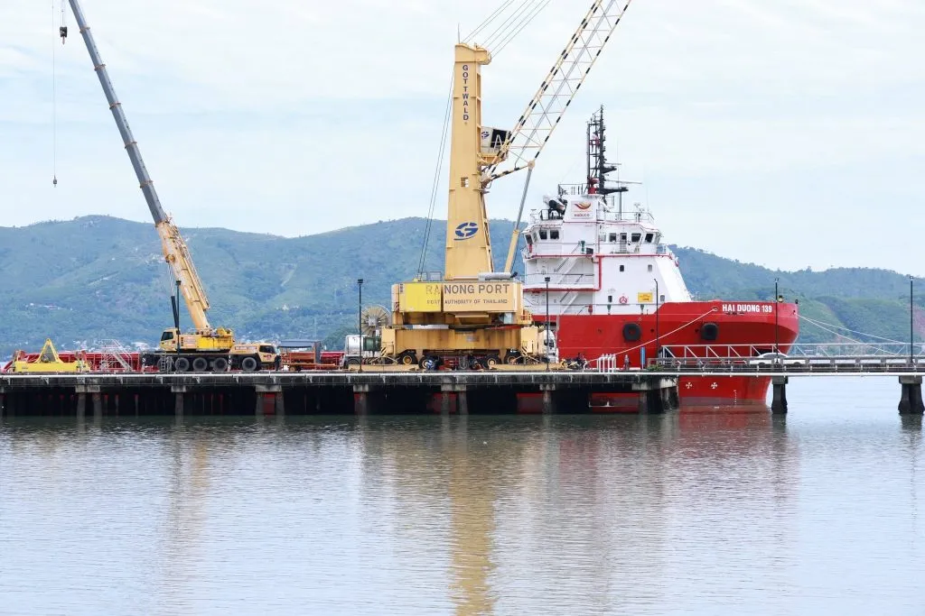 A red and white cargo ship docked at a pier, flanked by yellow industrial cranes.