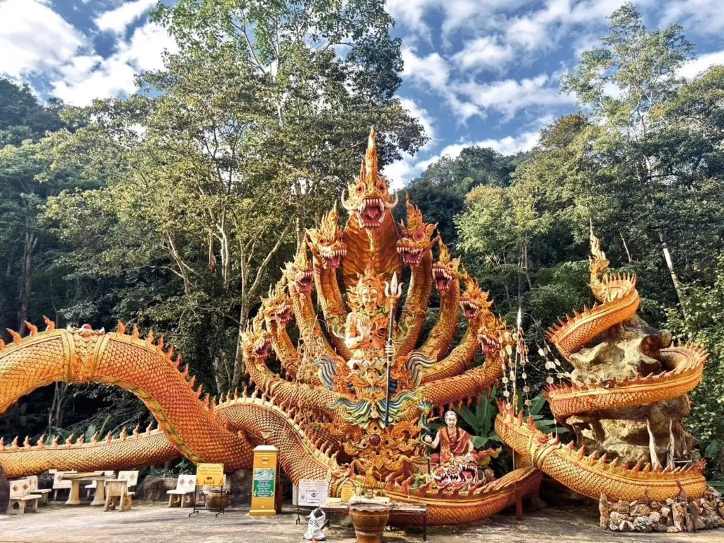 A large, golden, multi-headed Naga sculpture stands before a lush, green forest, framed by intricate, ornate Thai architectural details.