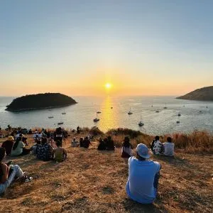 A crowd sits on a grassy cliff at sunset, watching small boats on the calm, sparkling sea below them.