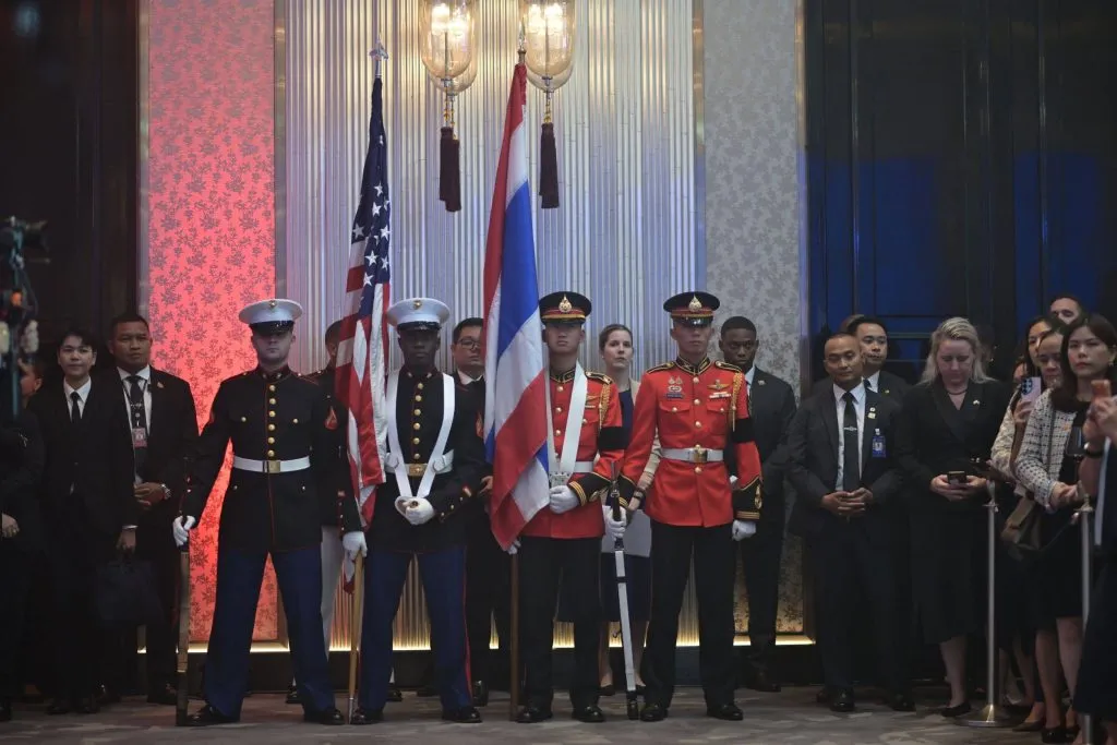Military color guard members holding American and Thai flags stand at attention indoors before a crowd.