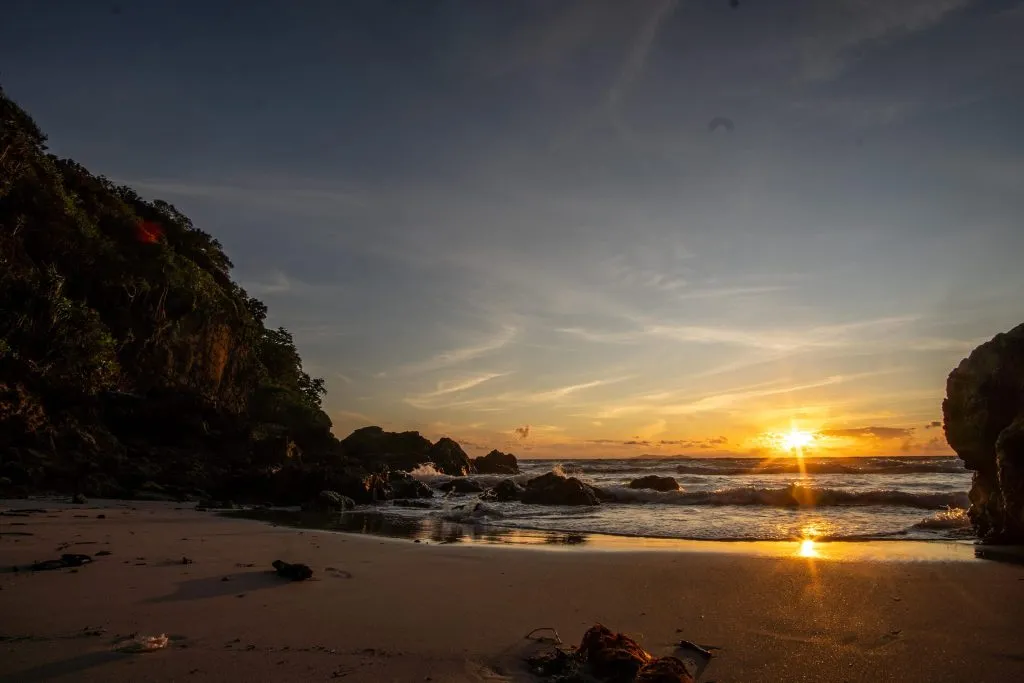 Waves gently lap onto a sandy beach at sunset, framed by dark rock formations and silhouettes of lush trees.