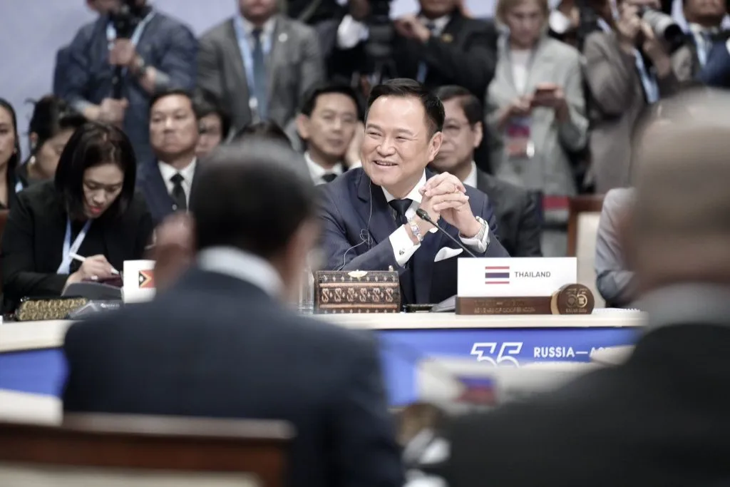 A smiling man in a suit sits behind a Thailand nameplate at a conference.