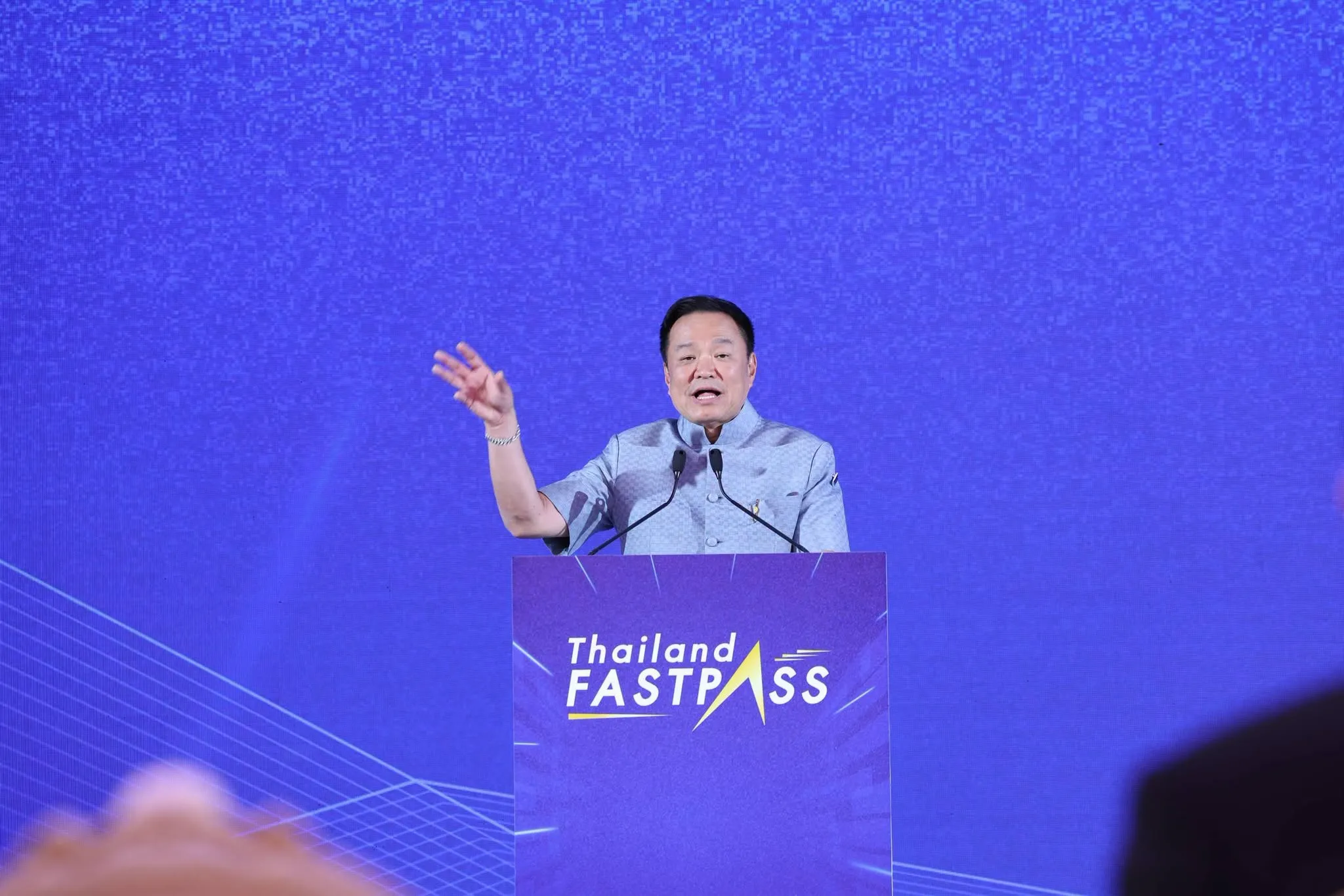 Prime Minister Anutin Charnvirakul stands at a purple "Thailand FASTPASS" podium, gesturing while speaking against a blue backdrop.