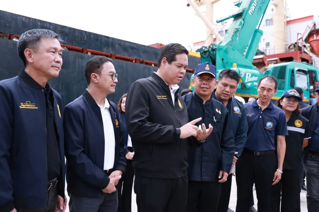 A group of Thai officials in dark jackets standing outdoors, discussing near industrial port machinery.