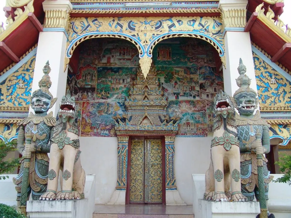 A colorful, ornate temple entrance features intricate murals and two tall, detailed guardian statues standing on either side, flanking doors.