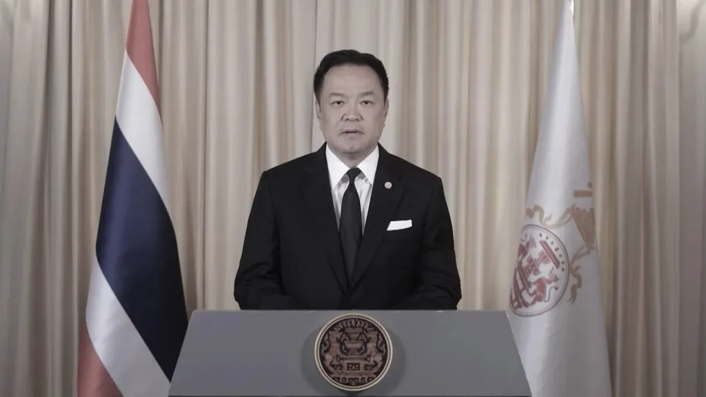 A man in a black suit speaks behind a podium between two flags.