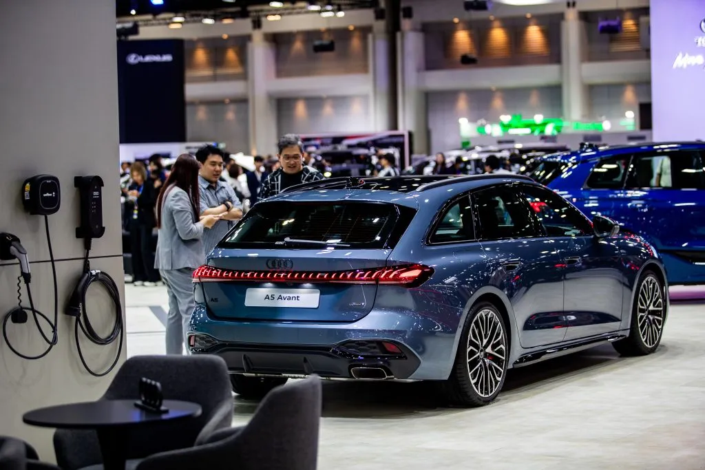 Attendees observe a dark blue Audi A5 Avant station wagon positioned near wall-mounted electric vehicle chargers.