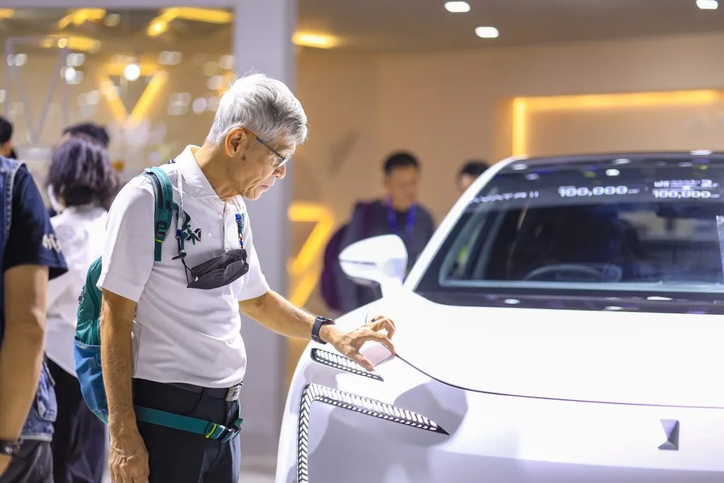 An elderly gentleman with a backpack examines the unique front LED headlight assembly of a white vehicle.