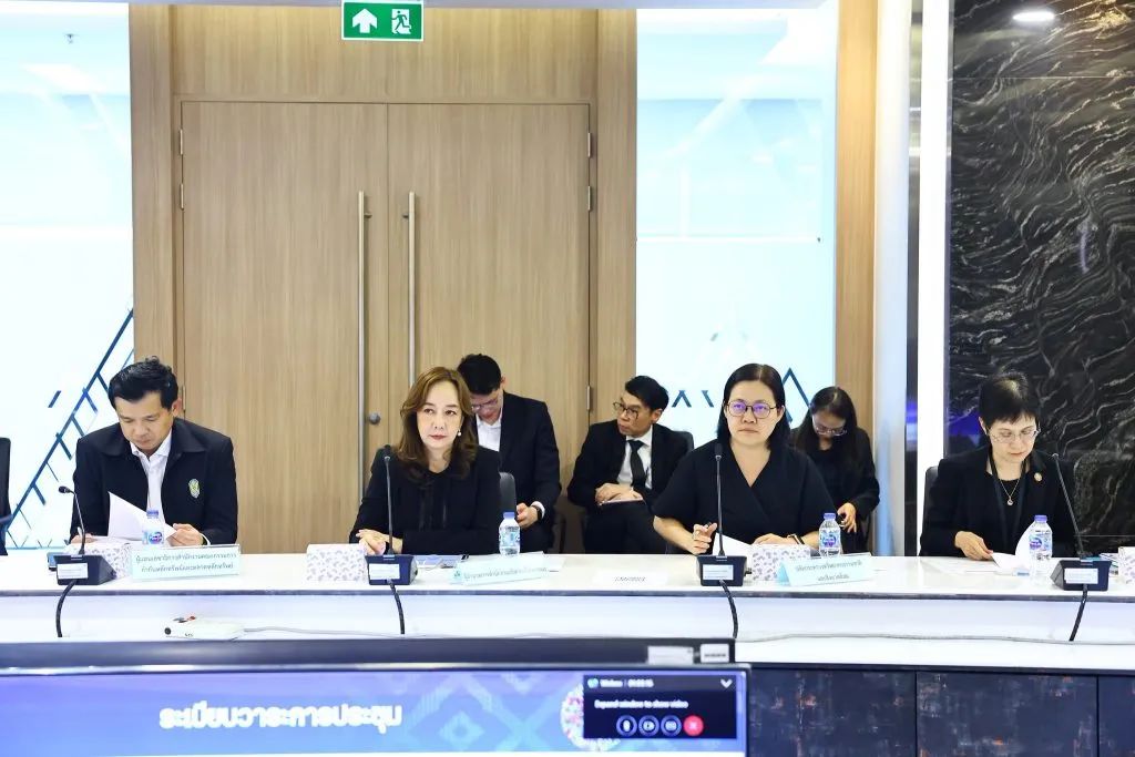 Four officials sit at a conference table with nameplates under an exit sign indoors.