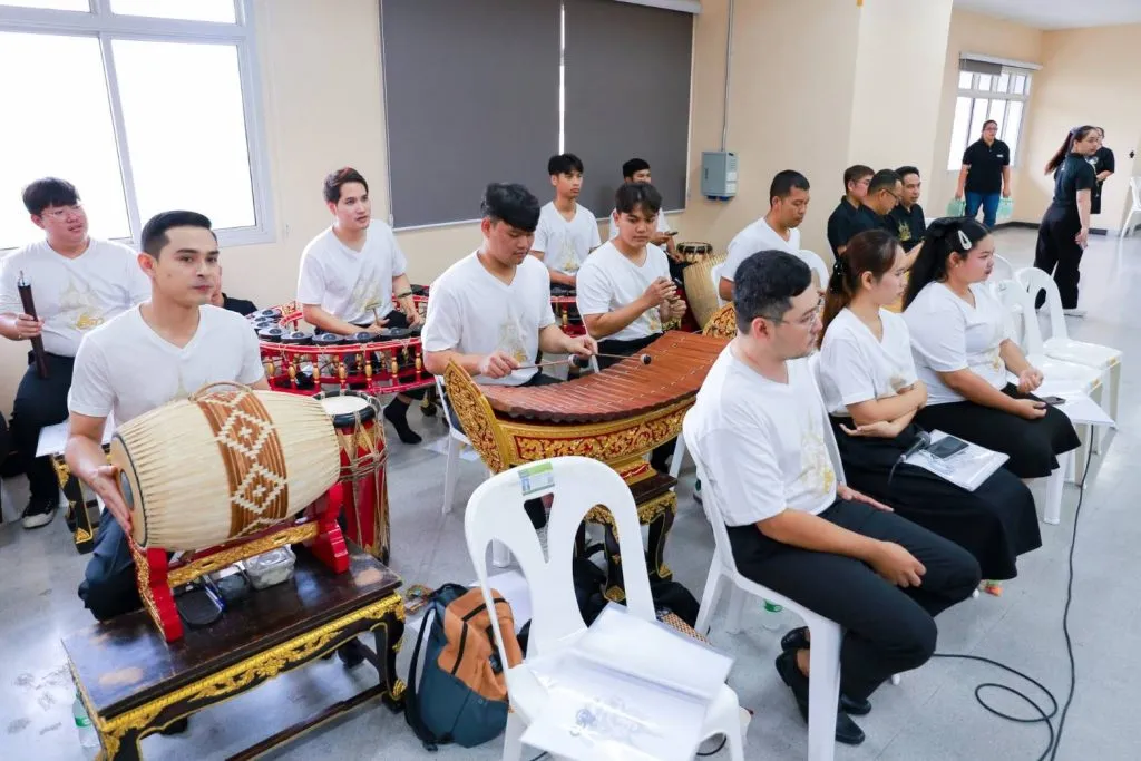 Musicians sit playing traditional Thai instruments, including drums and xylophones, inside a practice hall.