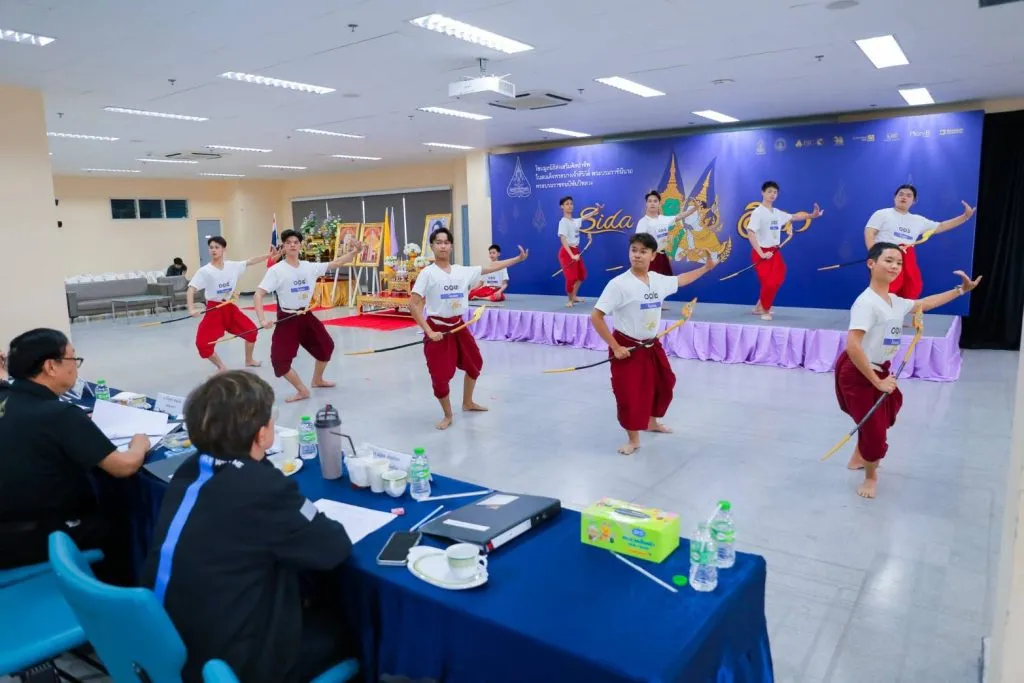 Young performers in white shirts and red traditional trousers perform Khon dance audition movements before seated judges in a hall.
