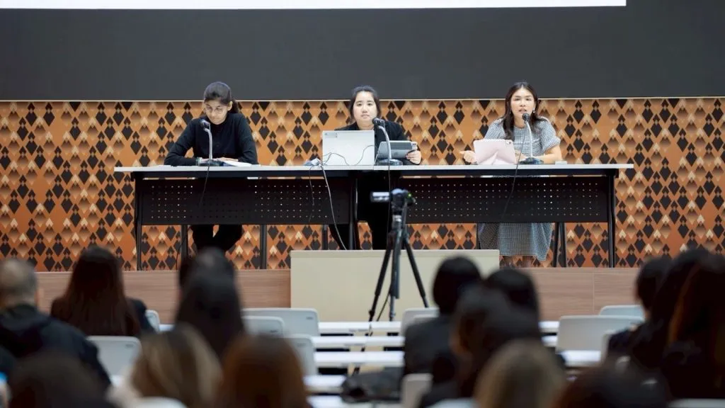 Three women sit panelist-style at a long elevated desk, addressing an audience in a lecture hall.
