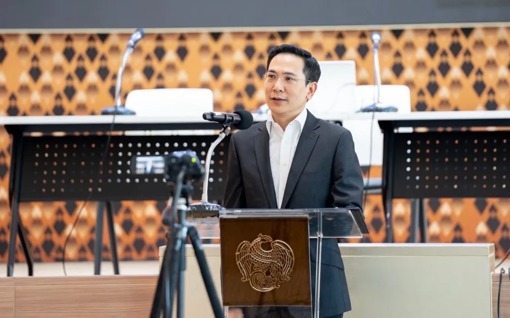 A man wearing glasses and a dark suit speaks confidently at a podium with microphones.