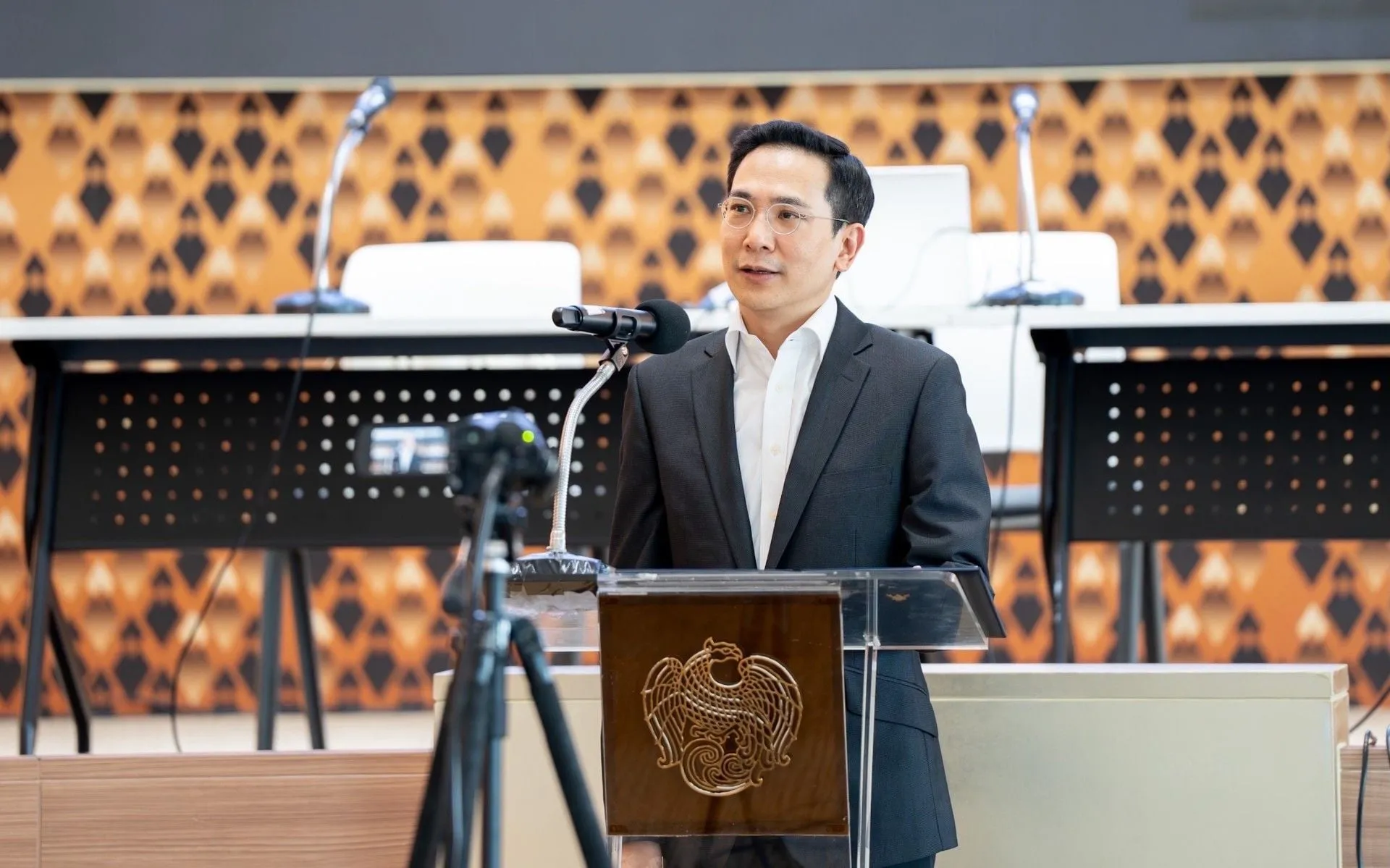 A man wearing glasses and a dark suit speaks confidently at a podium with microphones.