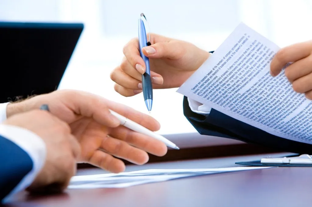 Close-up of corporate professionals holding pens and reviewing paperwork together at an office desk during a meeting.